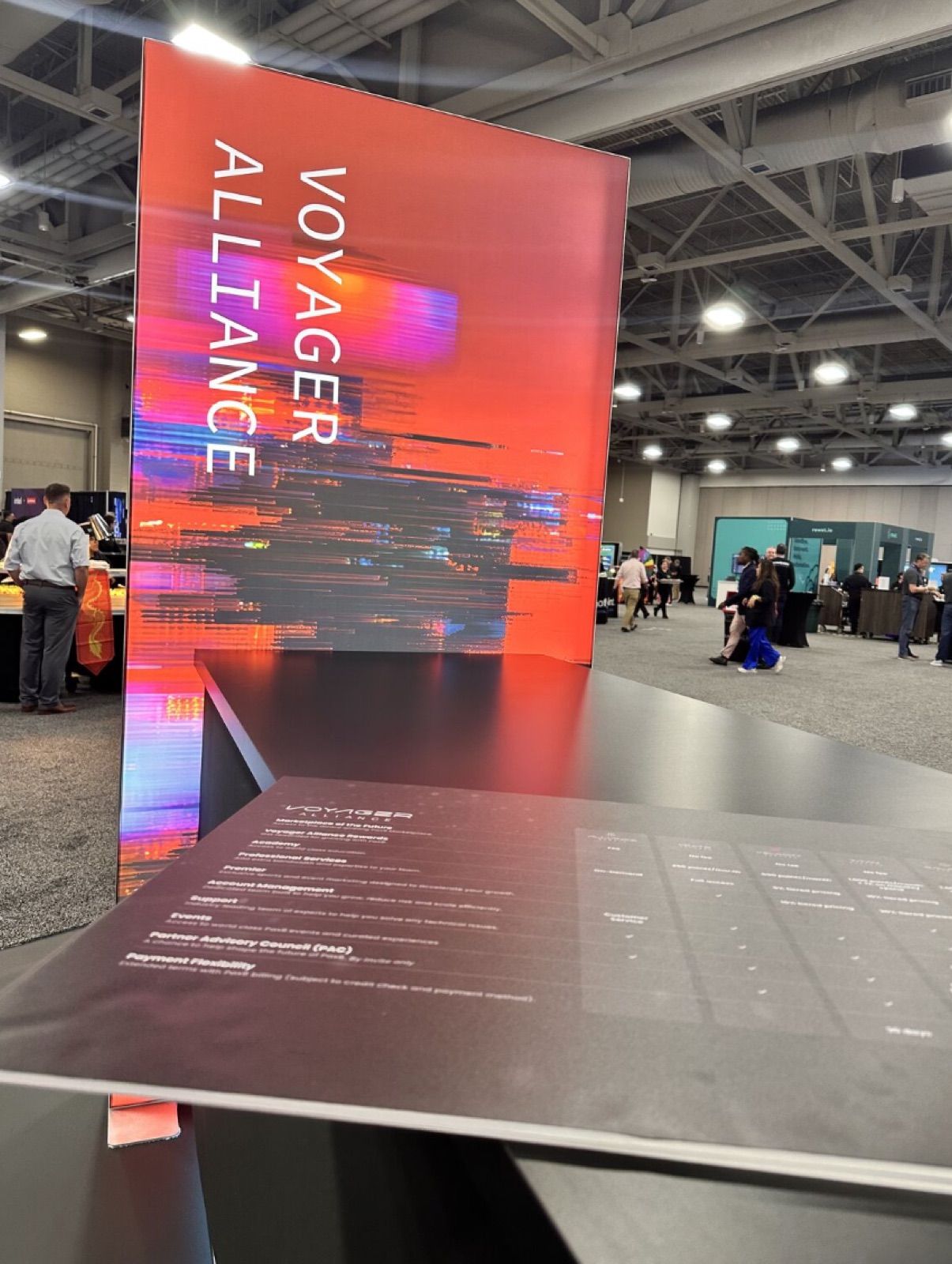 A tall Voyager Alliance sign at a booth, with a printed table of tier benefits in the foreground.